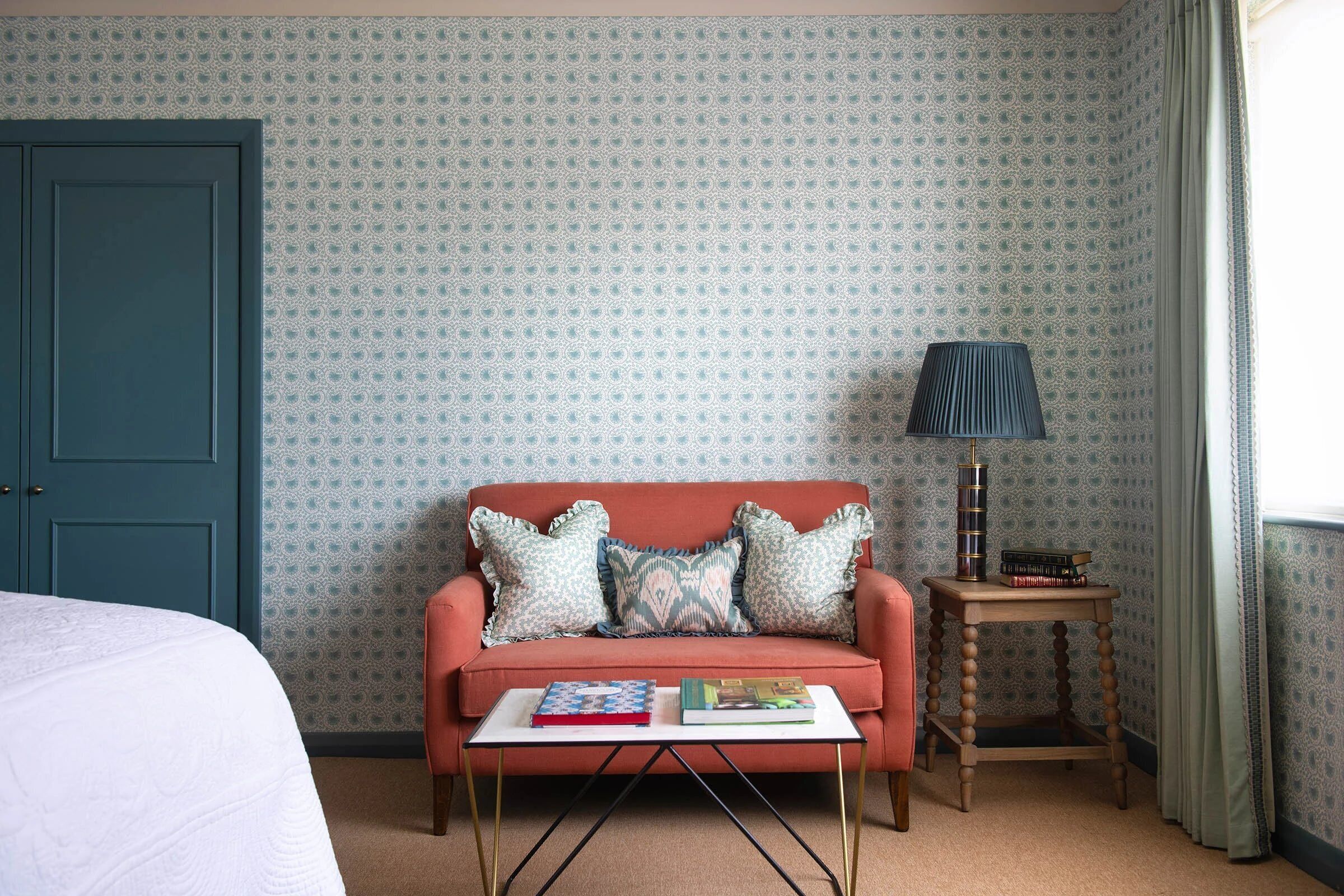 Una habitación con puerta color turquesa, papel tapiz estampado, un sofá rosa con cojines decorativos, una pequeña mesita de madera con una lámpara y libros, y una cama blanca con colcha.