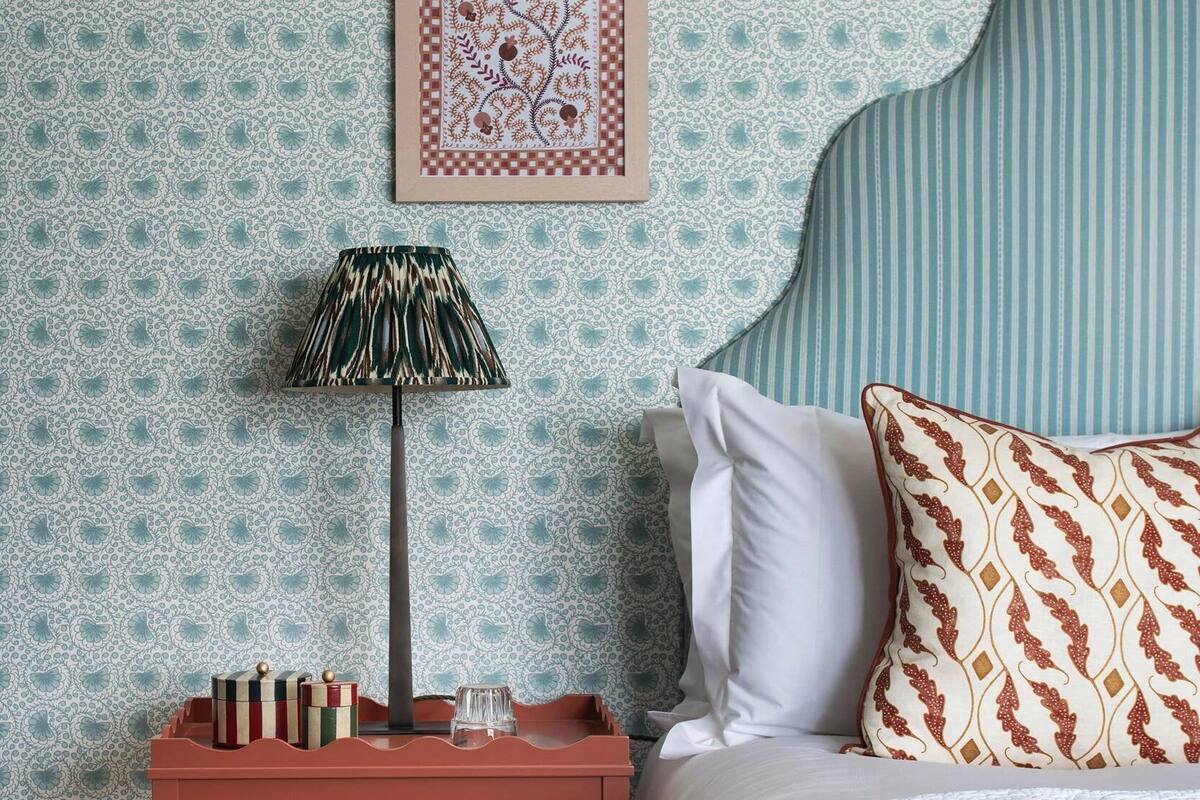 A neatly arranged bedroom with a patterned wallpaper, a framed artwork above the bed, a bedside lamp, and a small wooden nightstand with books and a glass.