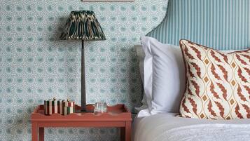 Quarto arrumado com papel de parede estampado, uma obra de arte emoldurada acima da cama, abajur e uma pequena mesa de cabeceira de madeira com livros e um copo.