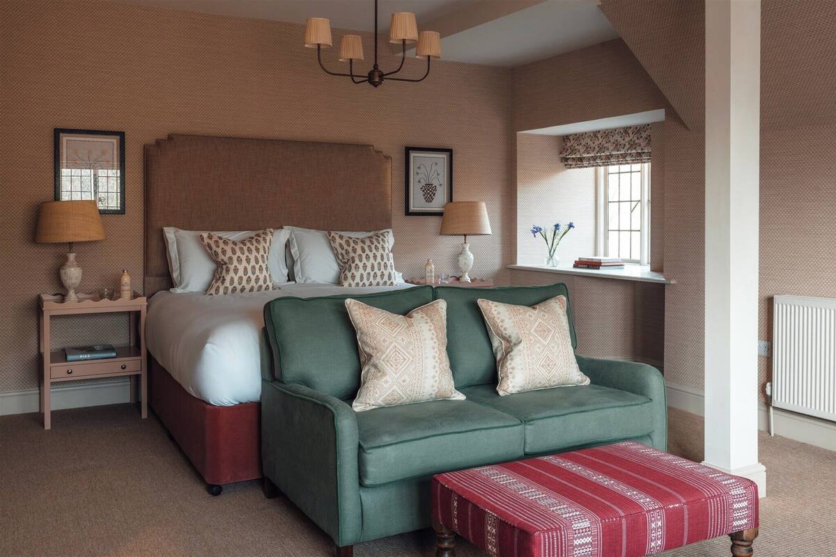 A bedroom with a large bed, a green sofa, a red ottoman, and a nightstand with lamps.