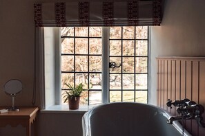 A bathroom with a bathtub, a window with a view of trees, a potted plant, and a mirror on a wooden table.