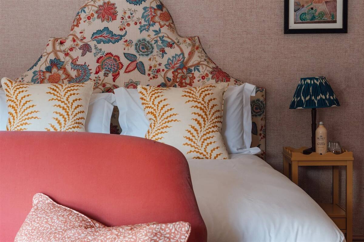 A bed with a floral headboard, a wooden nightstand with a bottle and glasses, and a patterned pillow.