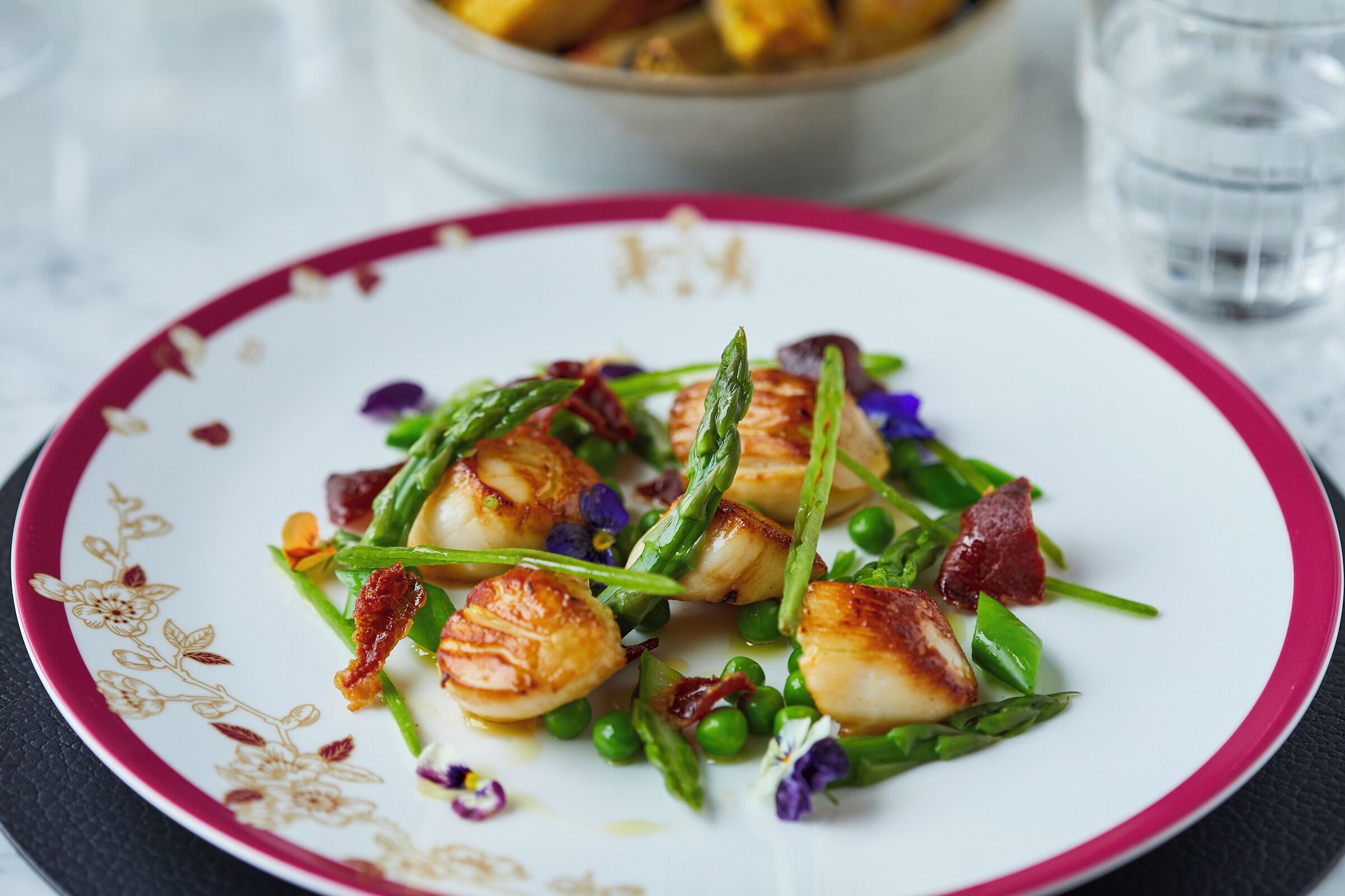 Une assiette de noix de Saint-Jacques poêlées, servies avec des asperges et des petits pois, et garnie de fleurs comestibles et de morceaux de lardons.