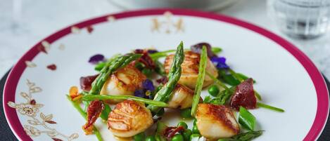 Une assiette de noix de Saint-Jacques poêlées, servies avec des asperges et des petits pois, et garnie de fleurs comestibles et de morceaux de lardons.