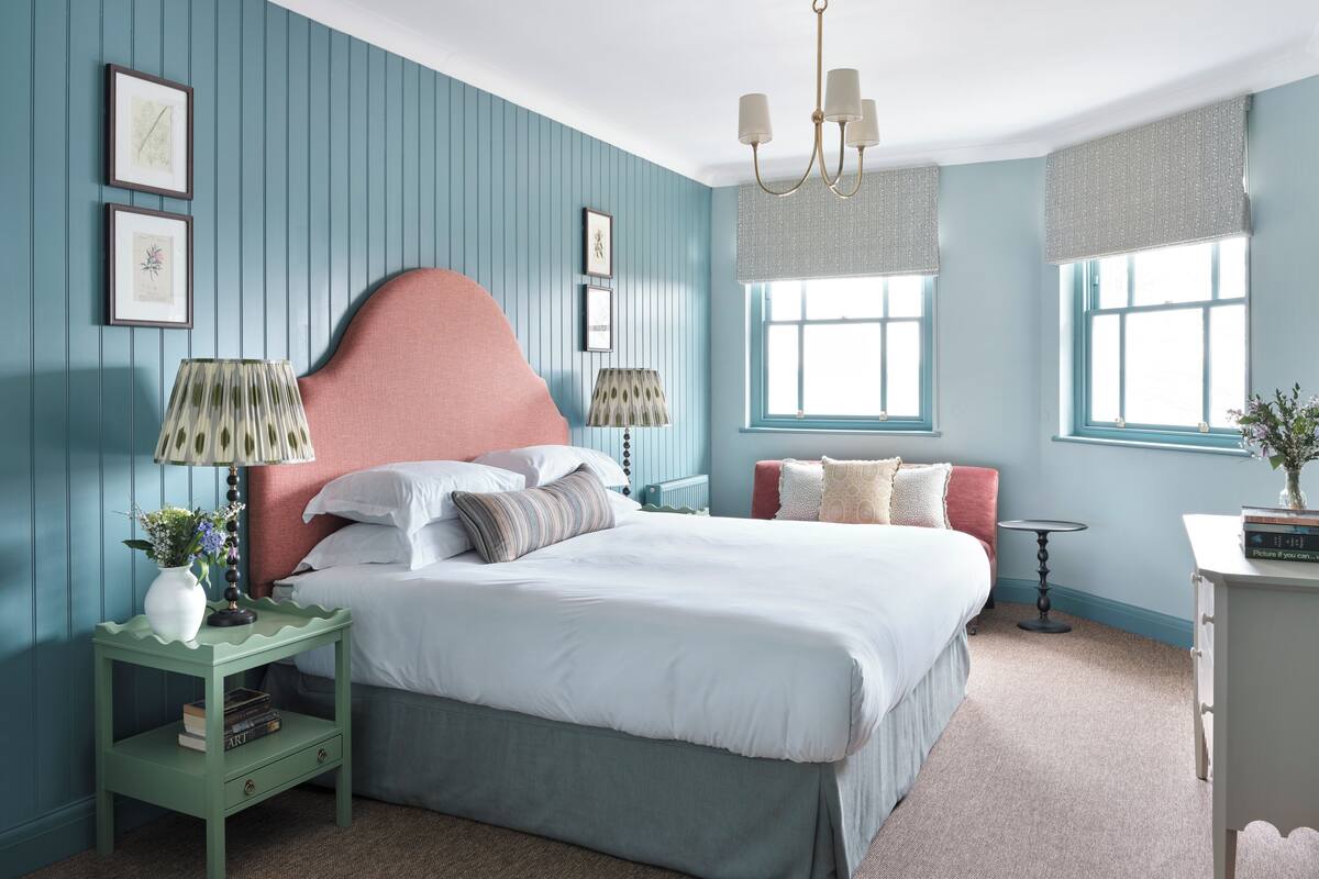 A bedroom with a large bed, two bedside tables, a nightstand, a chandelier, and a window with curtains.