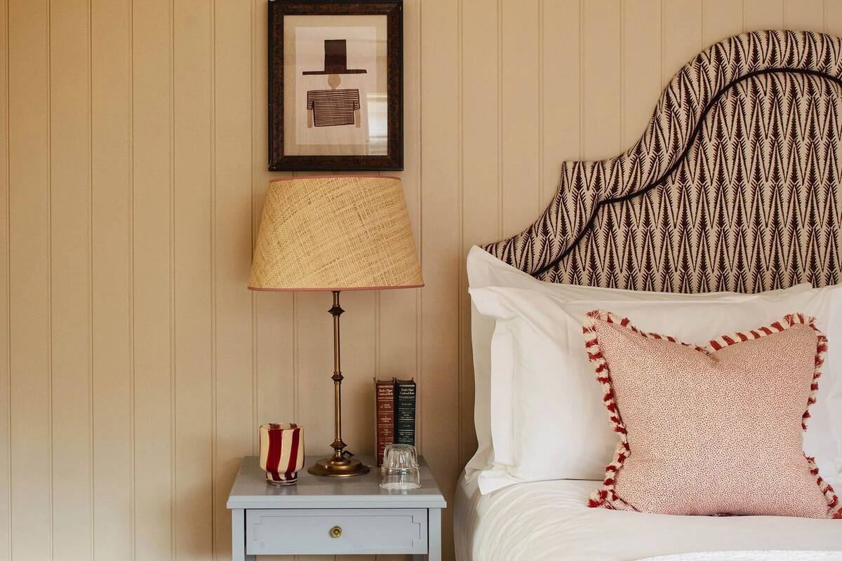 A neatly arranged bedroom with a bed, a bedside table with a lamp, a framed picture, and a bookshelf.