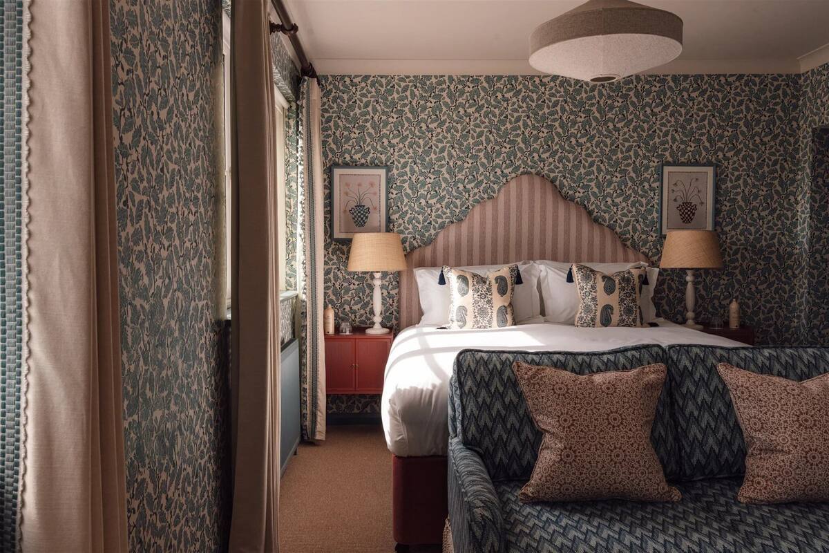 A hotel room with a bed, two bedside lamps, a nightstand, a patterned sofa, and a large window with curtains.