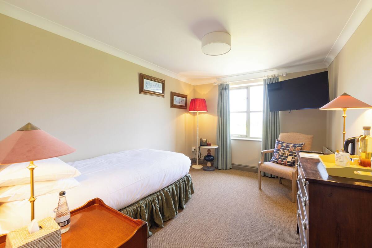 A hotel room with a bed, bedside lamps, a chair, a desk with a lamp, a TV, and a window with curtains.