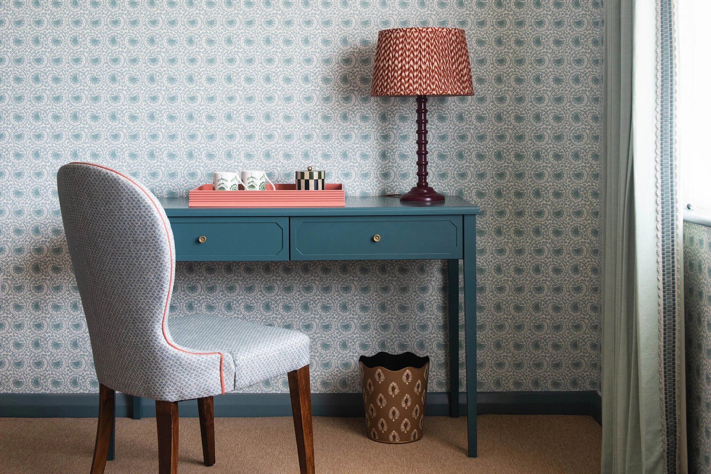 A room with a patterned wallpaper, a desk with drawers, a chair, a lamp, and a trash bin.