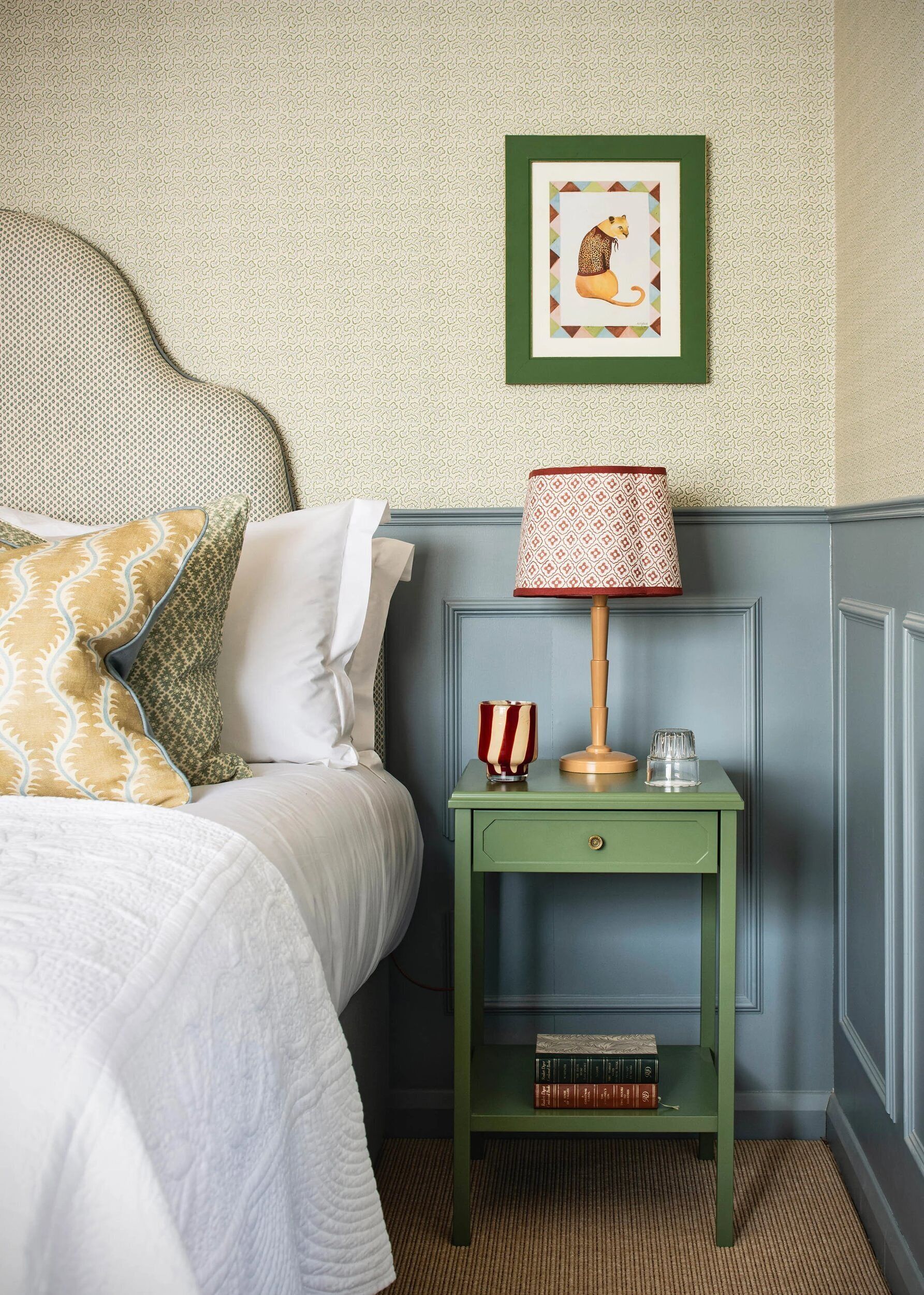 A neatly arranged bedroom with a bed, pillows, a bedside table with a lamp, a framed picture, and a small shelf with books.