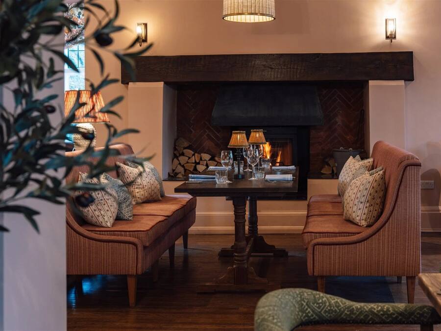 A cozy living room with a fireplace, two cushioned chairs, a table with glasses, and a hanging lamp.