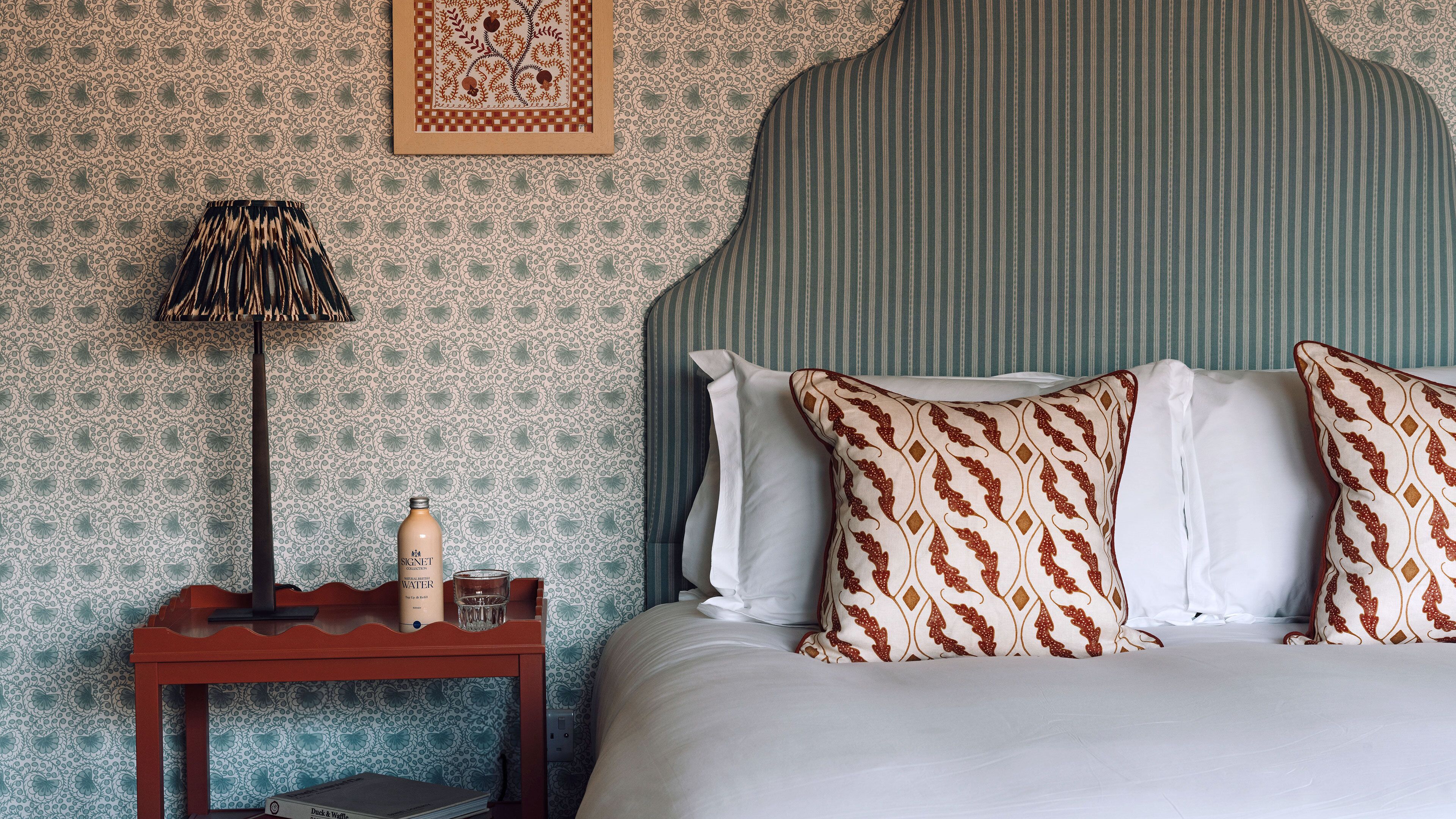 Un lit bien fait, agrémenté d’oreillers à motifs, une table de chevet avec une bouteille et une lampe, et un mur recouvert d’un papier peint à motifs floraux.