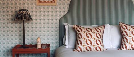 Un lit bien fait, agrémenté d’oreillers à motifs, une table de chevet avec une bouteille et une lampe, et un mur recouvert d’un papier peint à motifs floraux.