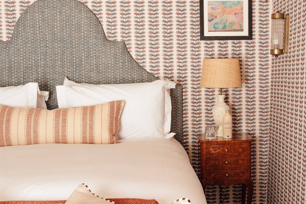 A neatly made bed with patterned pillows and a headboard, a bedside table with a lamp and a bottle, and a wall with a floral wallpaper pattern.