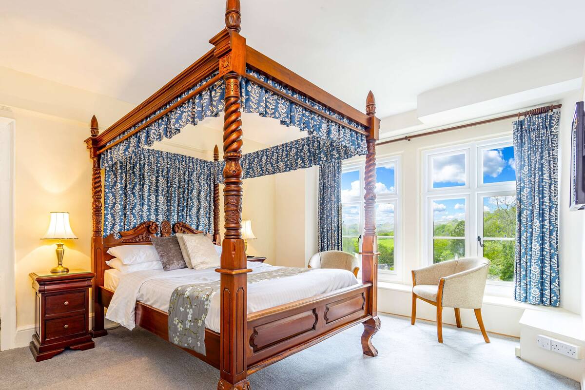 Un lit à baldaquin, une table de chevet avec une lampe, une chaise près de la fenêtre et une vue sur la verdure à l’extérieur.