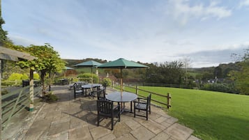 An outdoor patio with stone flooring, round tables, and chairs, surrounded by a wooden fence and a grassy lawn.