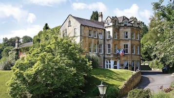 A large, historic building with multiple windows and a stone facade, surrounded by lush greenery and a stone wall.