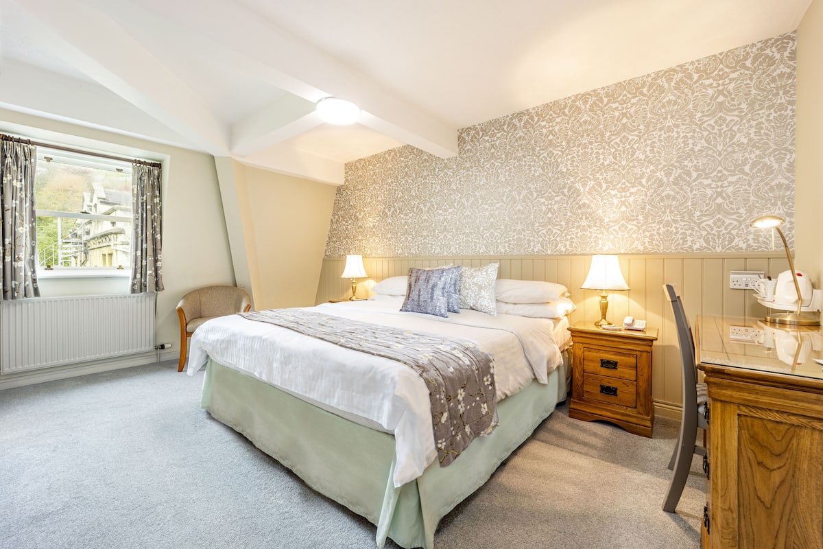 Une chambre à coucher avec un grand lit, deux tables de chevet avec des lampes, une chaise, un bureau et un papier peint à motifs.
