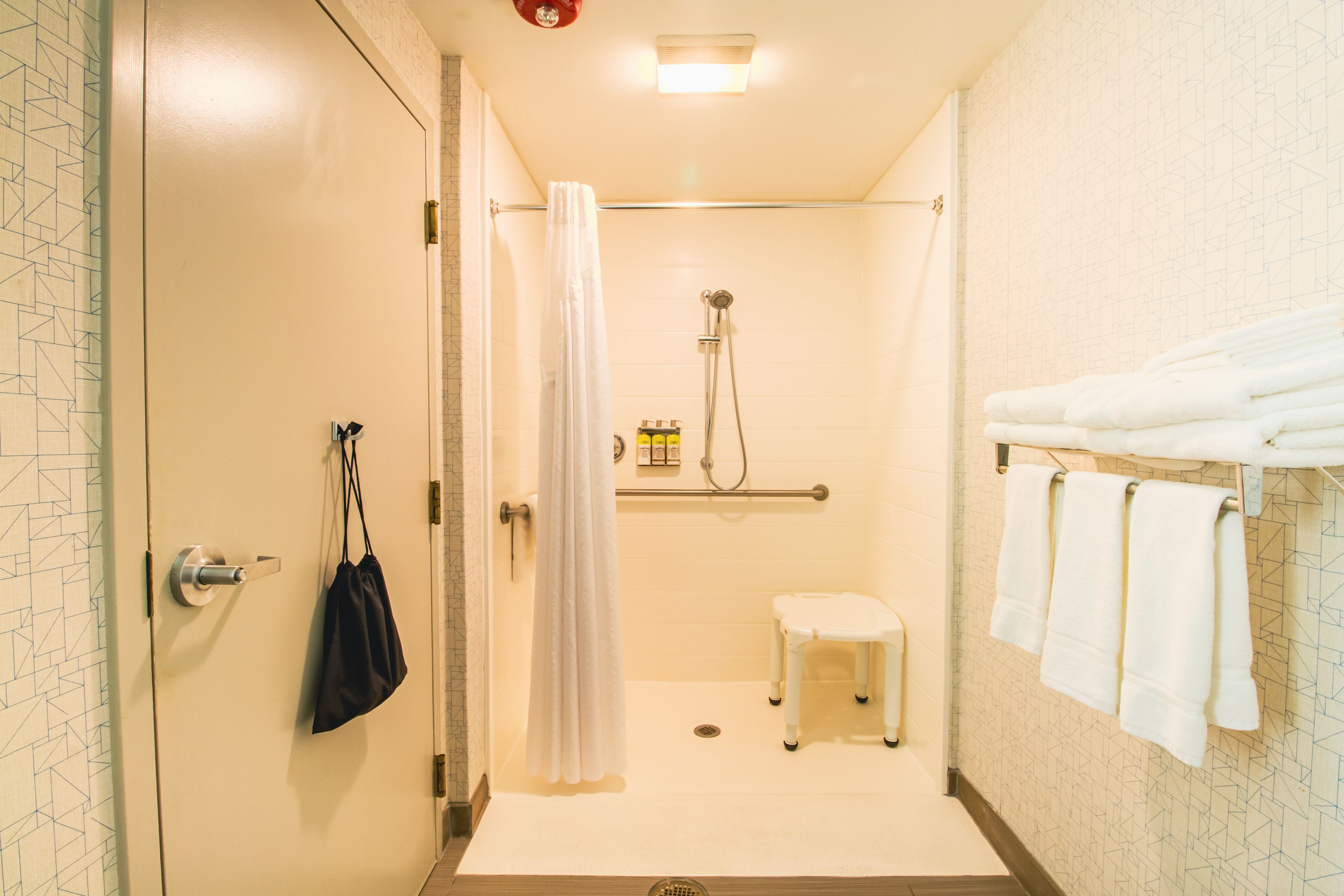 Et lille, lukket bruseområde med et hvidt brusehoved, et hvidt toilet og en lille, hvid skammel. Der er en bøjle med flere håndklæder og en sort taske hængende på væggen.