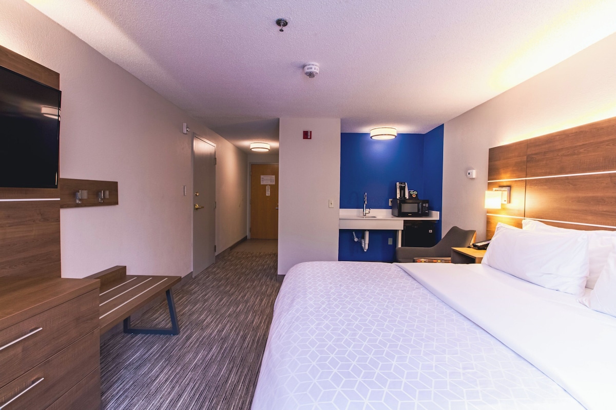 A hotel room with a large bed, a desk with a chair, a TV mounted on the wall, and a kitchenette area.