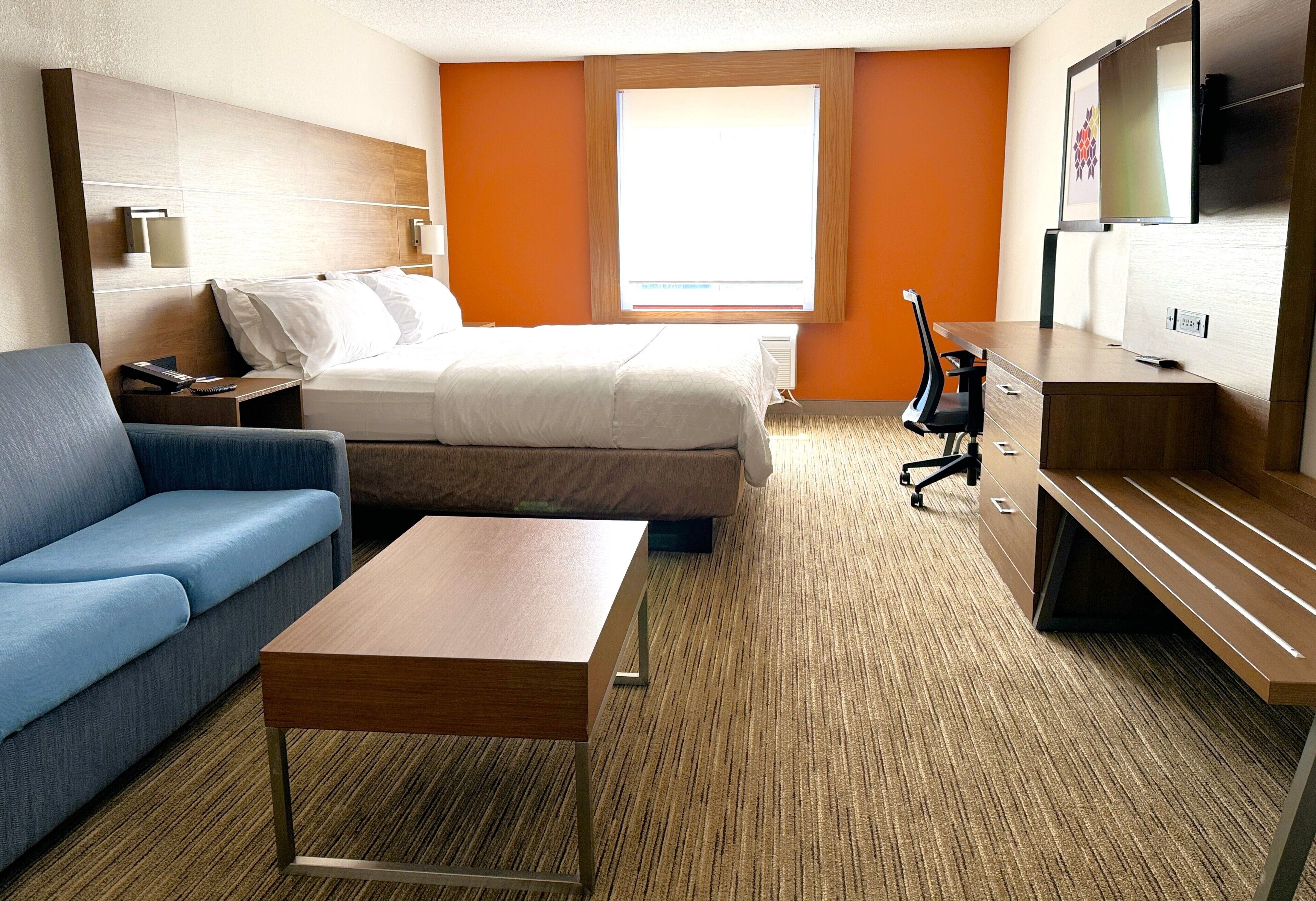 A hotel room with a bed, a blue sofa, a wooden coffee table, a desk with a chair, and a TV.