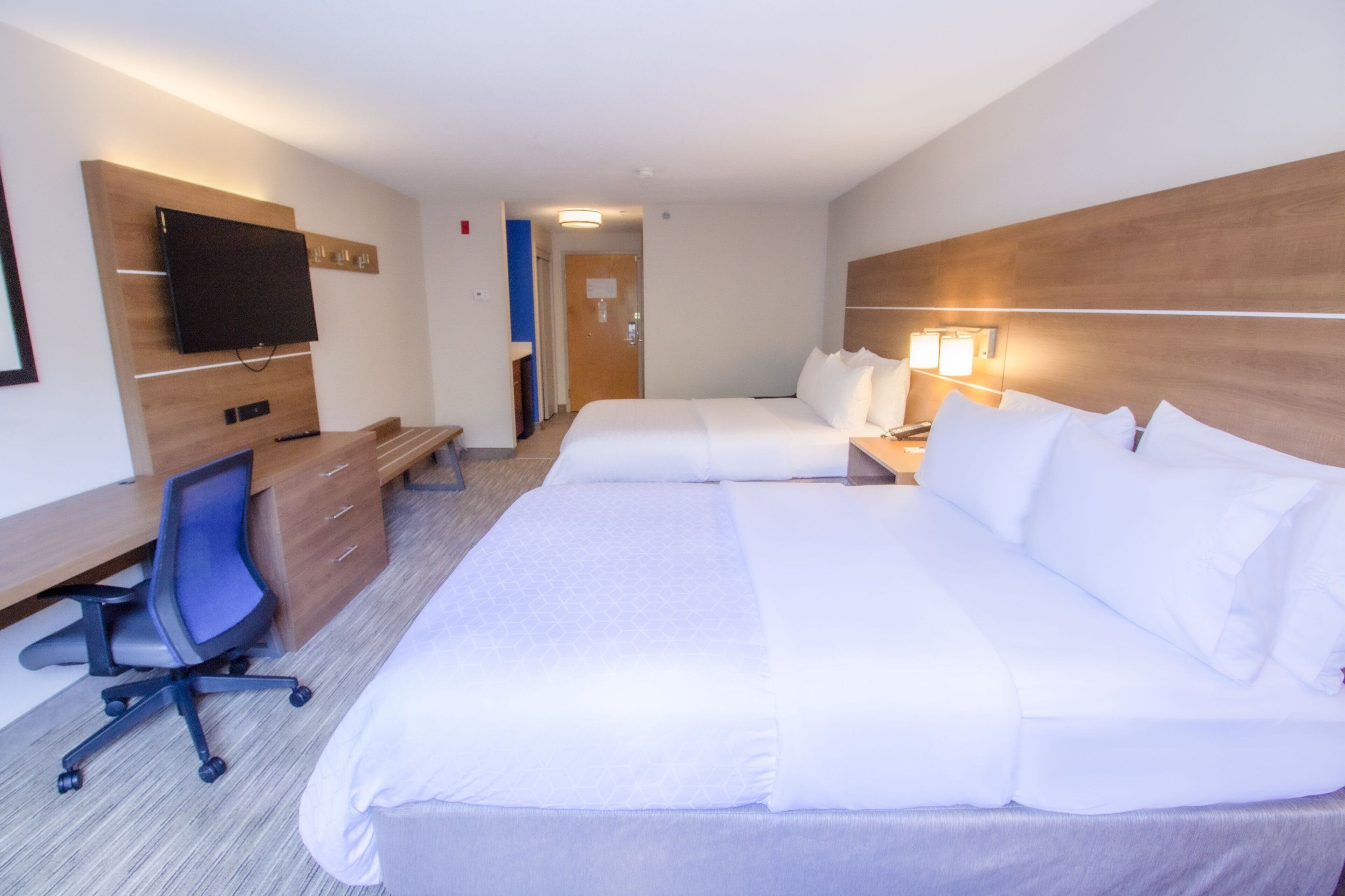 A hotel room with a large bed, a desk with a chair, a television mounted on the wall, and a bedside lamp.