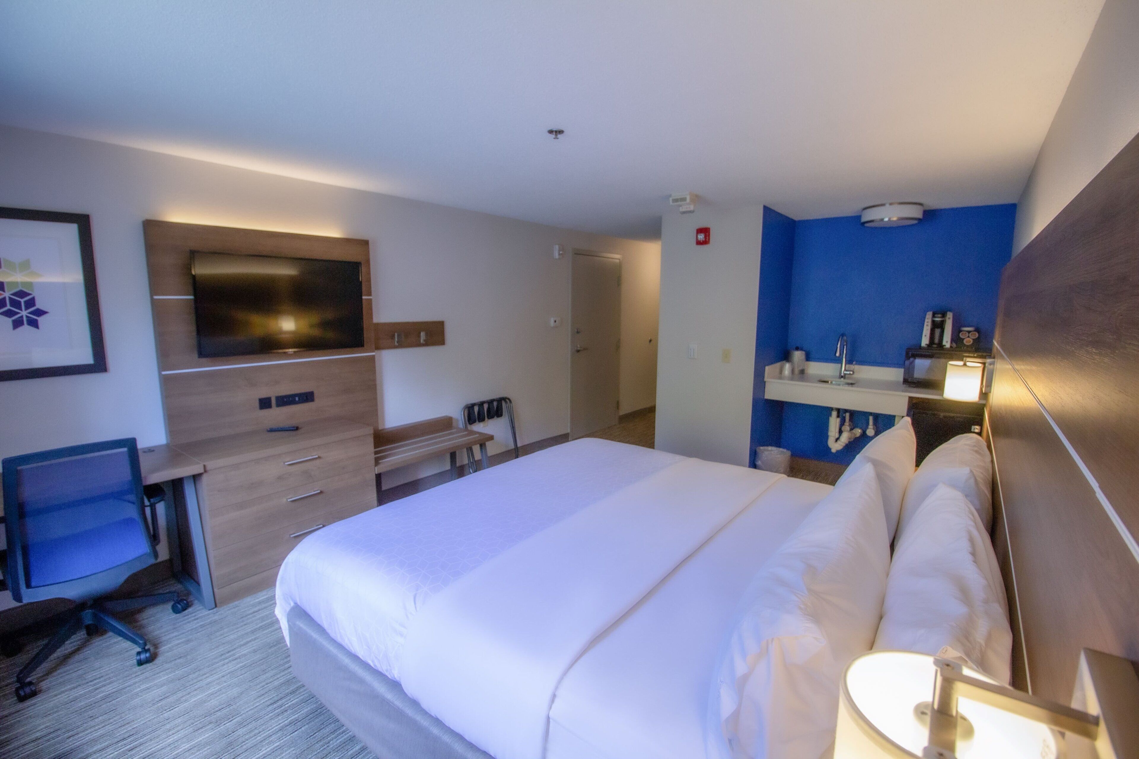 A hotel room with a large bed, a desk with a chair, a TV mounted on the wall, and a bathroom with a mirror and sink.