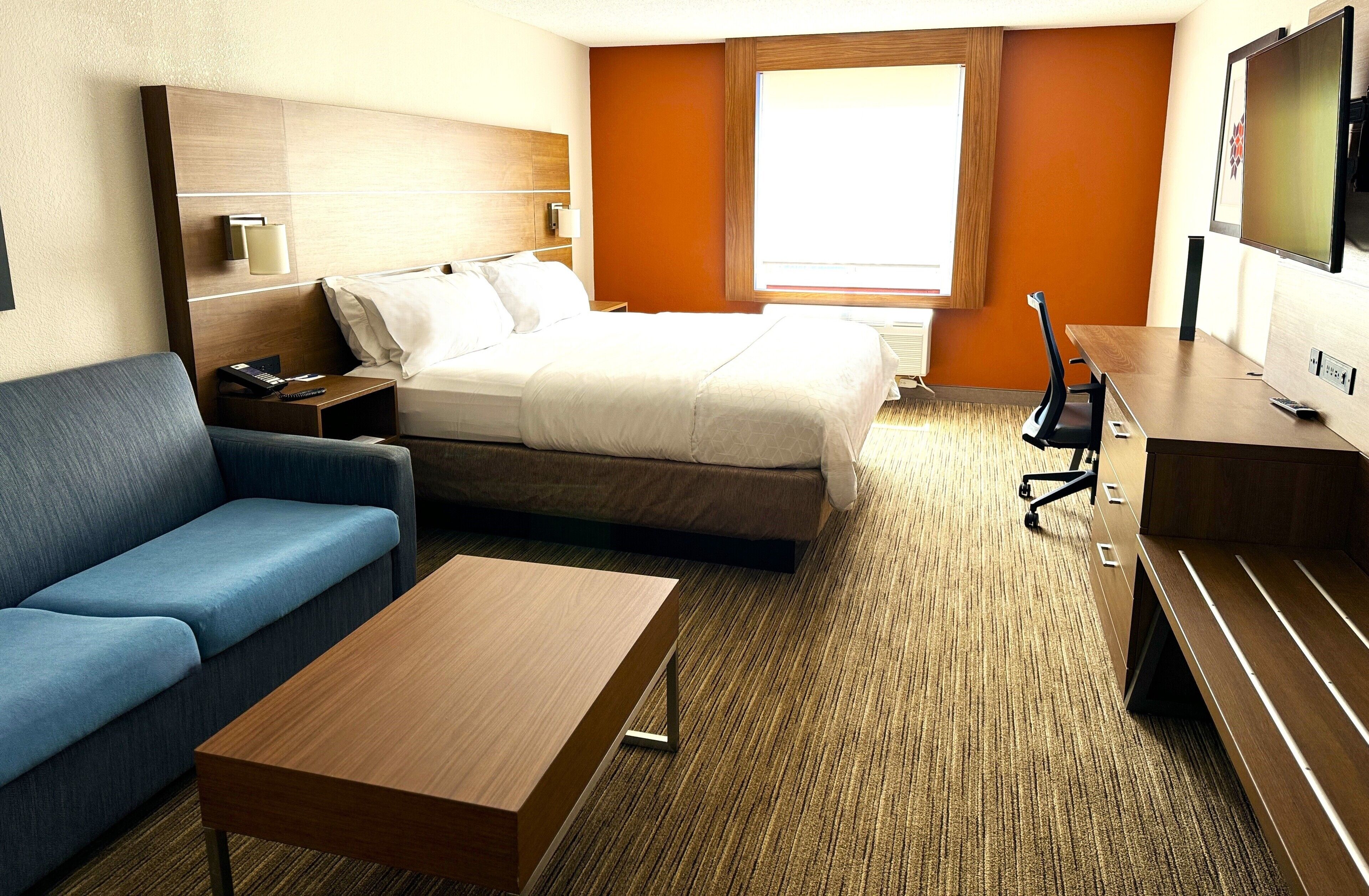 A hotel room with a bed, a blue sofa, a wooden coffee table, a desk with a chair, a TV, and a window with a view.