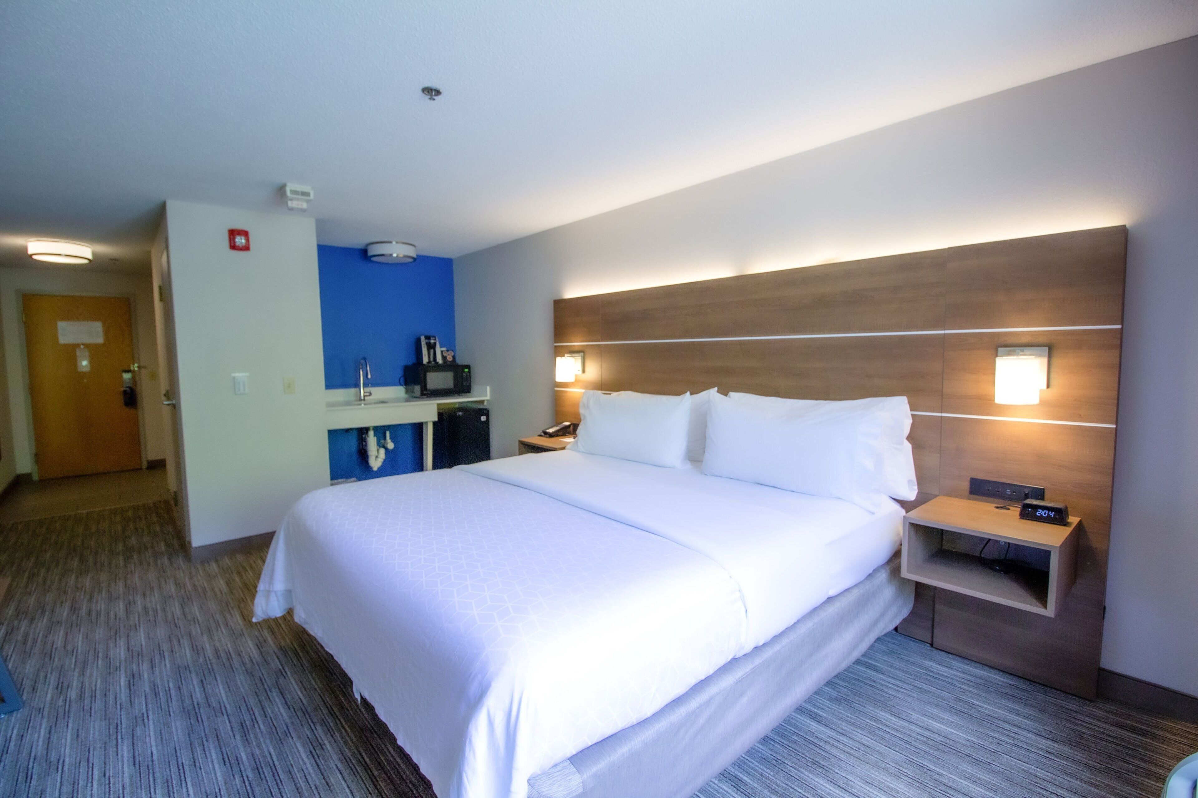 A hotel room with a large bed, a bedside table with a clock, a nightstand, a desk with a computer, and a blue accent wall.