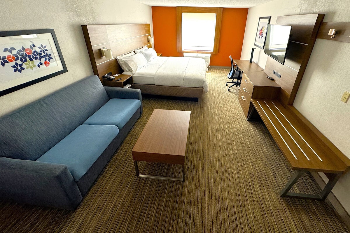 Ein Hotelzimmer mit einem Bett, einem blauen Sofa, einer hölzernen Bank, einem kleinen Tisch, einem Fernseher und einem gerahmten Bild an der Wand.