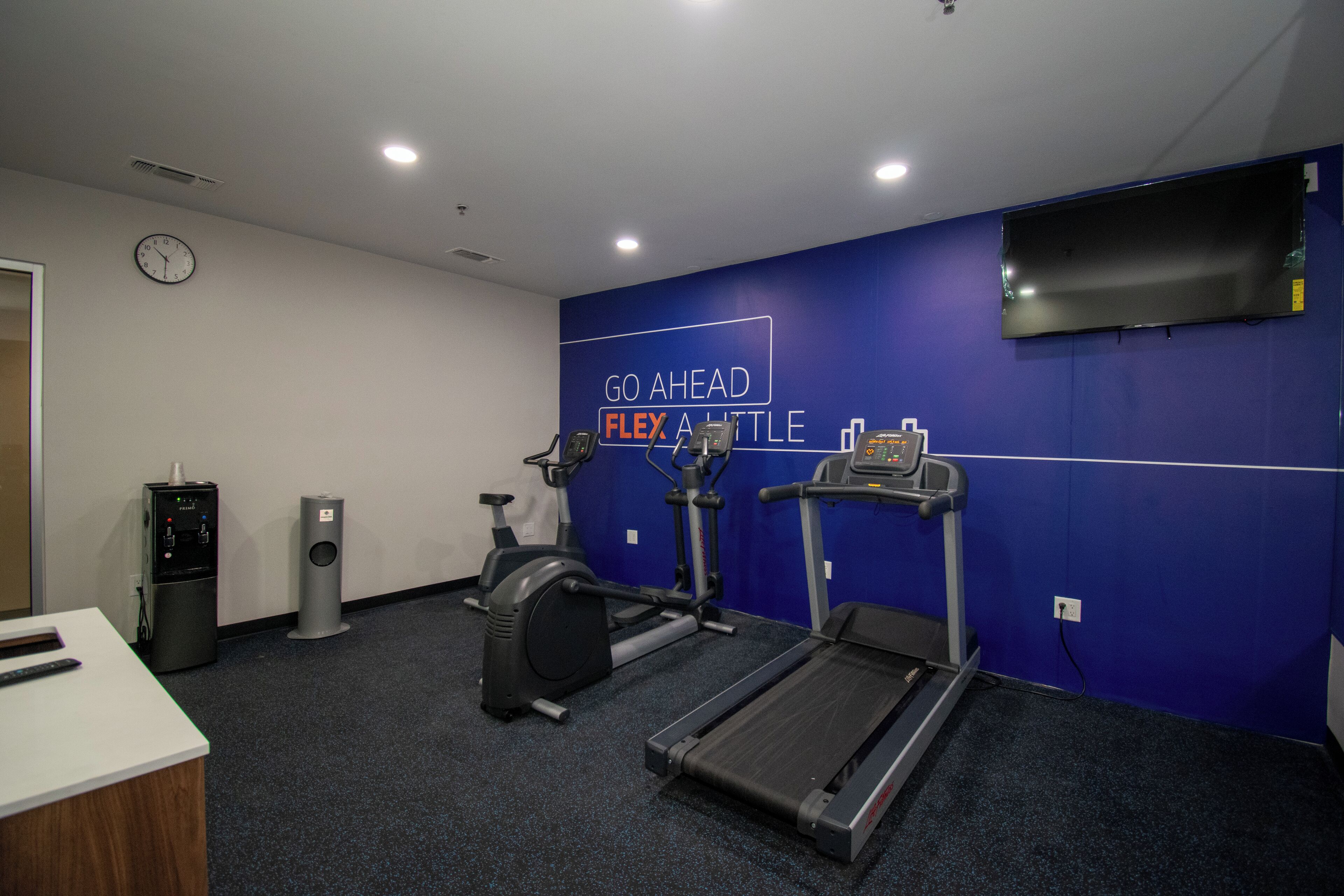 A gym with a treadmill, elliptical, and stationary bike, a water dispenser, and a clock on the wall.