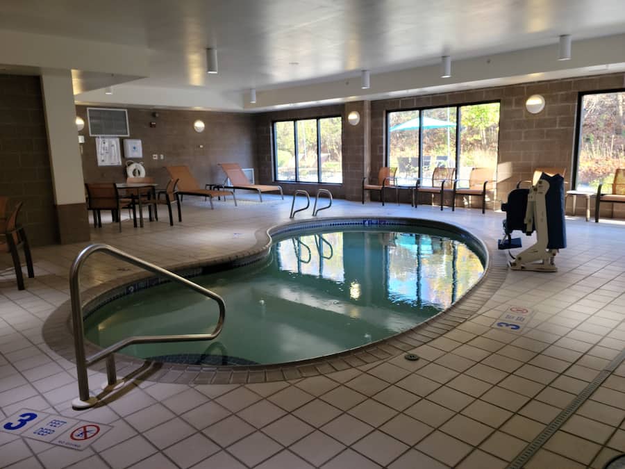 Una piscina interior con suelo de baldosas, sillones de descanso y vistas a árboles a través de grandes ventanales.
