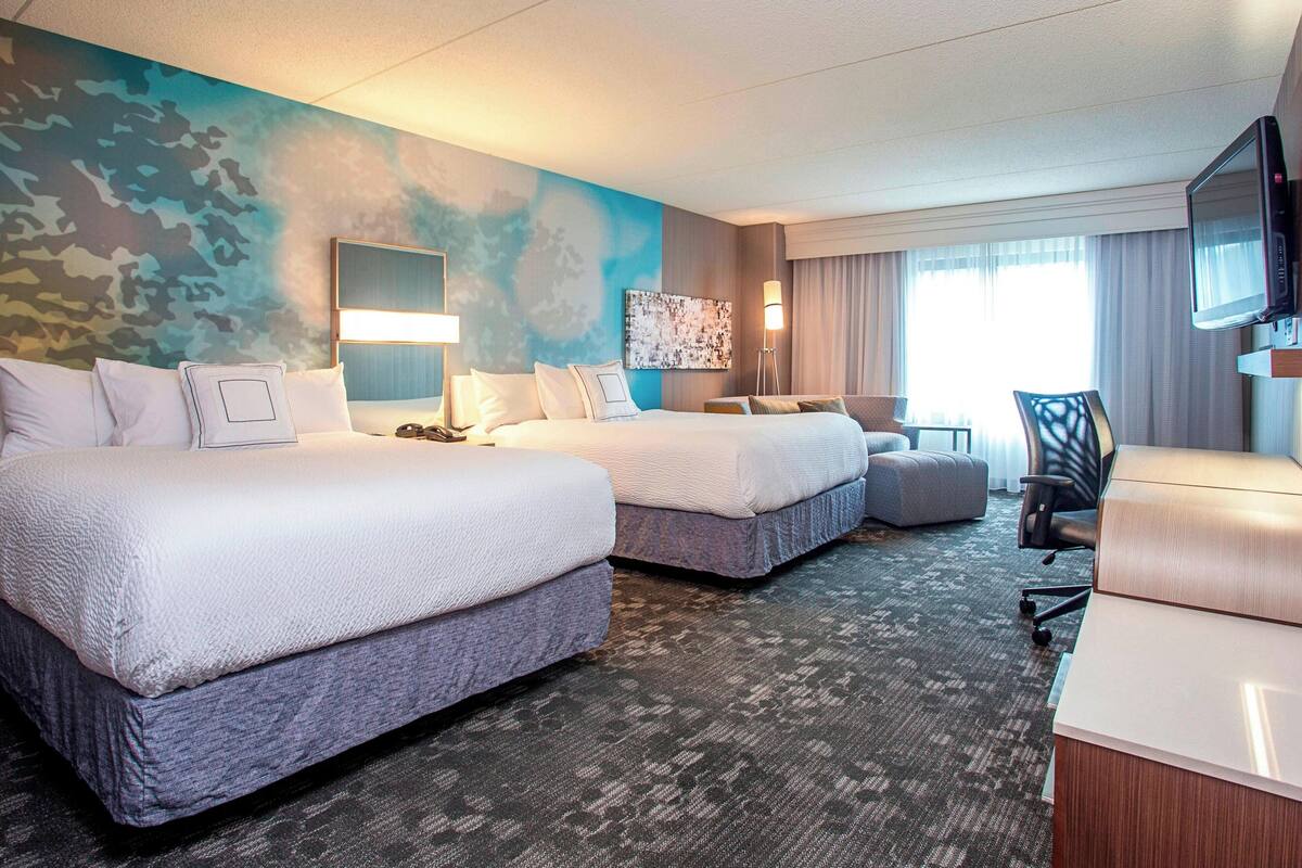 A hotel room with two beds, a desk, a chair, a TV, and a large window.