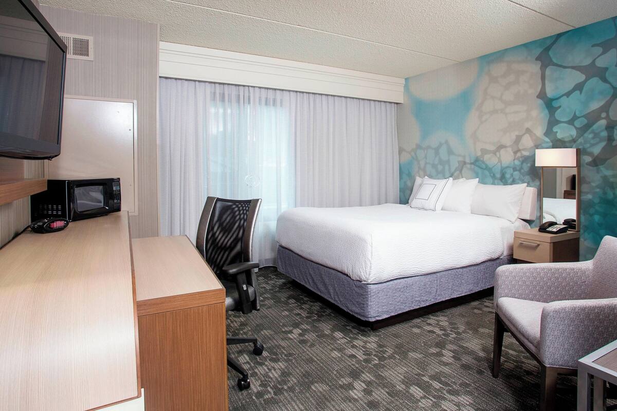 A hotel room with a bed, desk, chair, TV, and a painting on the wall.
