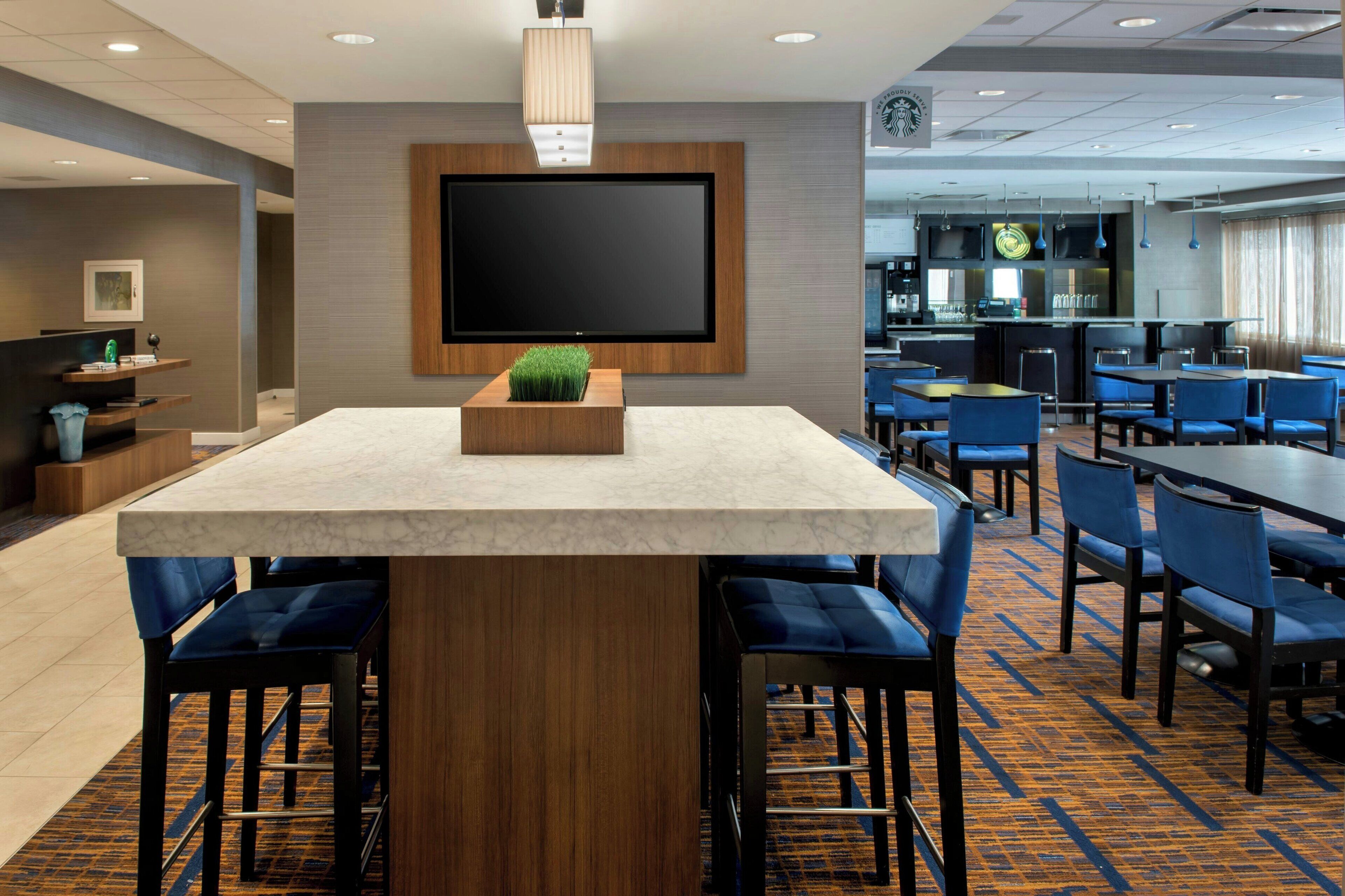 Una hall moderna di un hotel con un grande televisore a schermo piatto montato a parete, un’area bar con sedute blu e una moquette decorata.