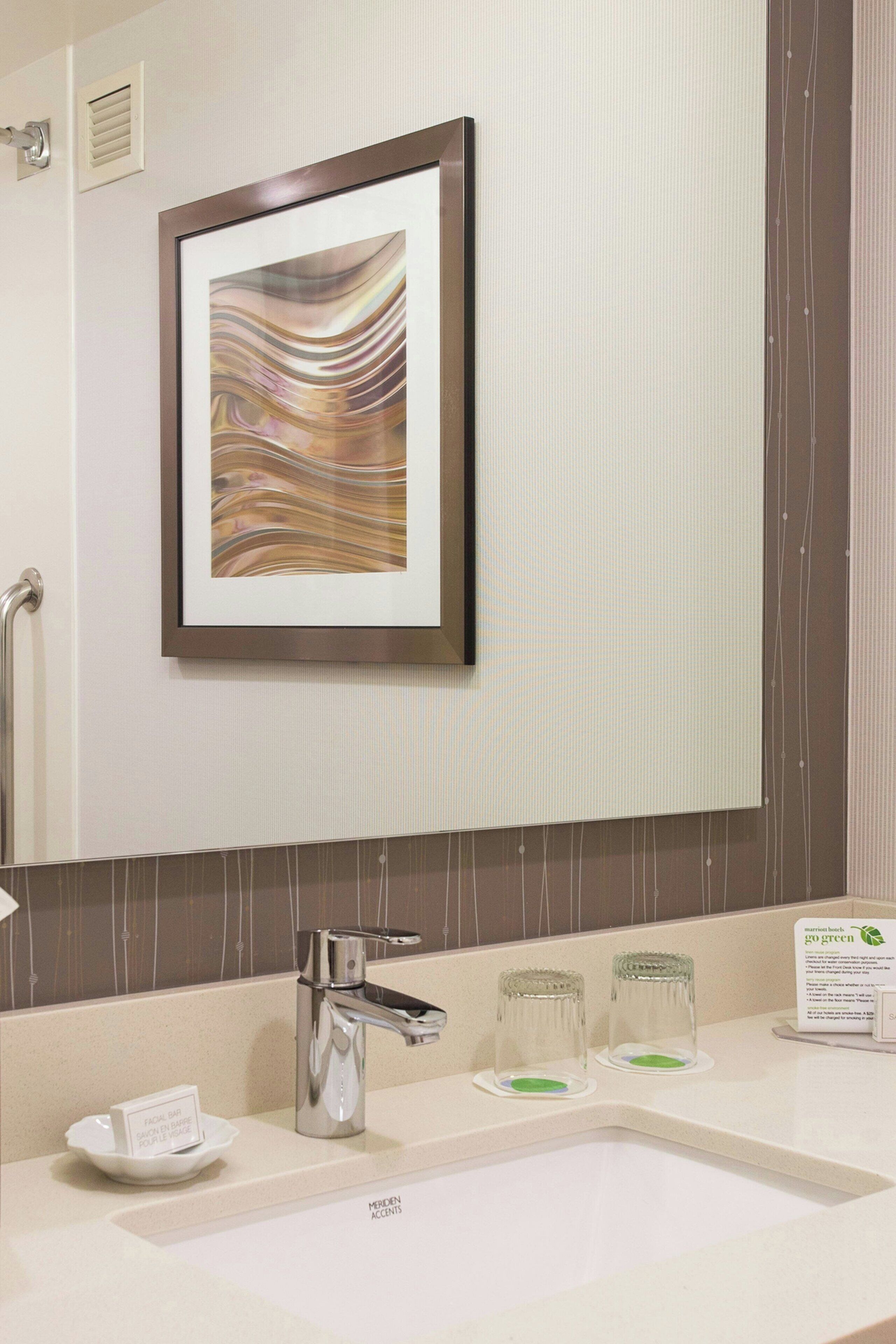 A bathroom with a large mirror, a framed abstract artwork, a modern faucet, and a sink with two glass soap dispensers.