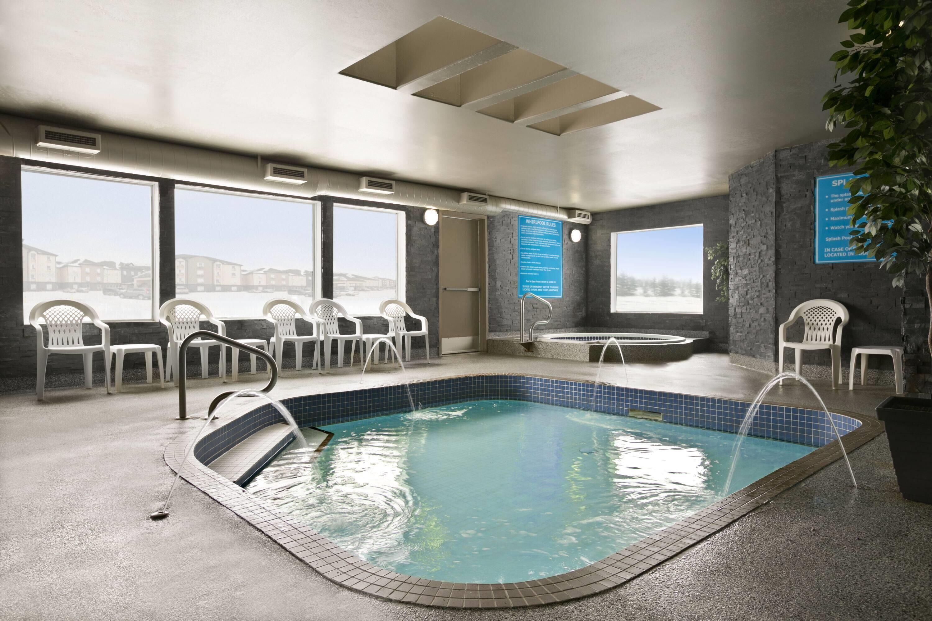 Una piscina interior con vistas amplias a través de grandes ventanales, sillas blancas de plástico y un área de spa.