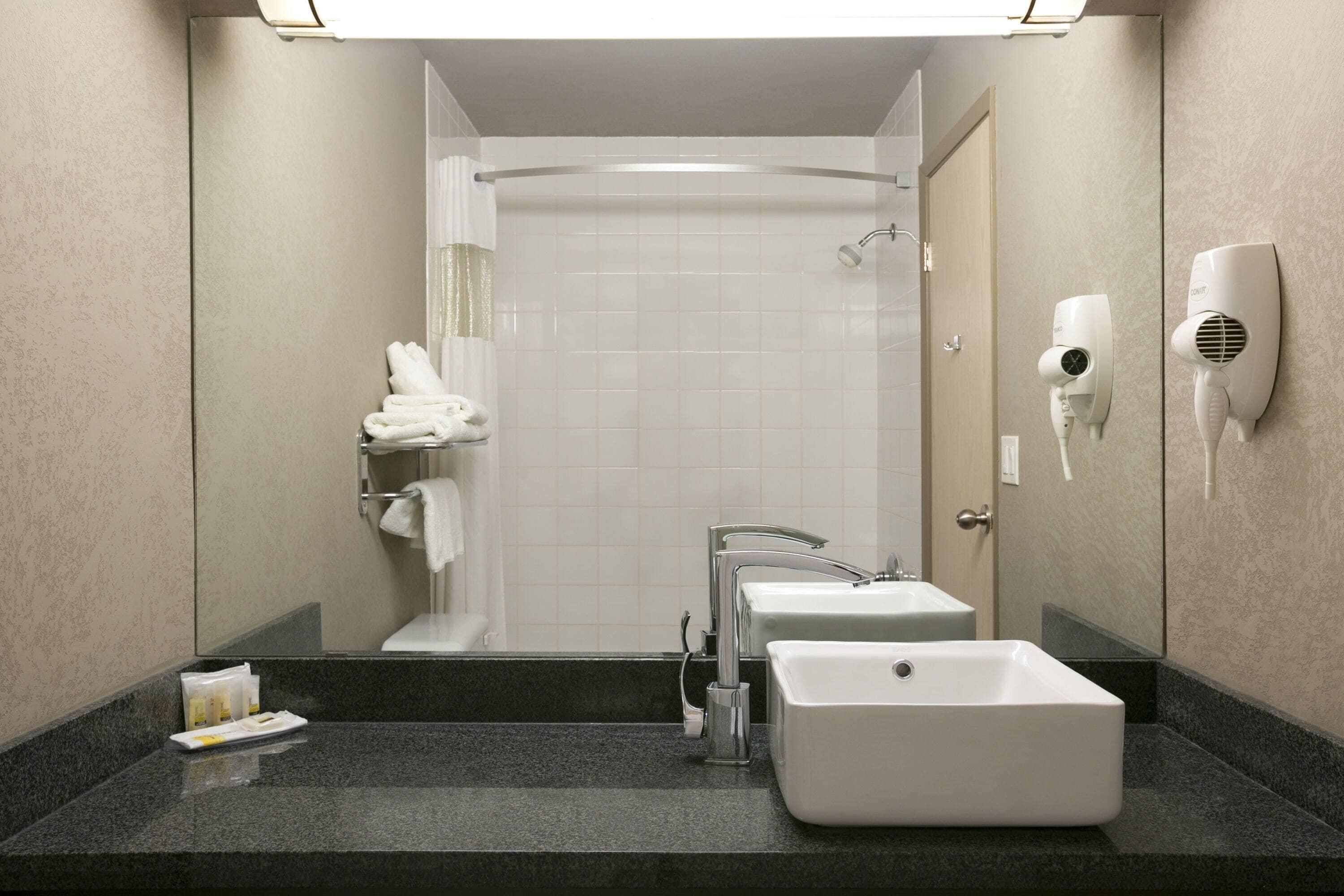 Une salle de bain moderne avec un grand miroir, un lavabo, une douche et deux sèche-cheveux fixés au mur.