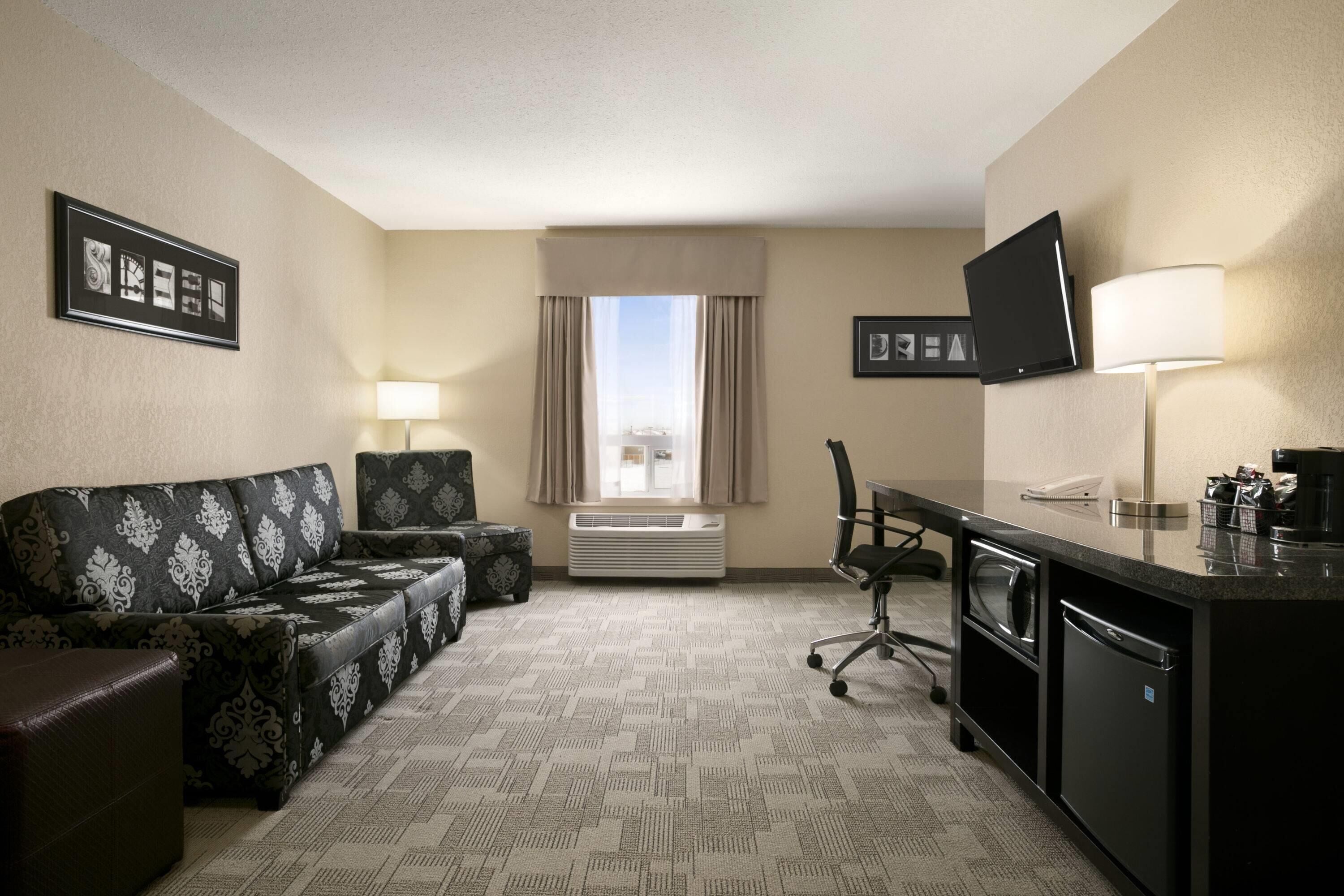 Habitación de hotel con sofá, escritorio, silla, televisión, lámpara y ventana con cortinas.