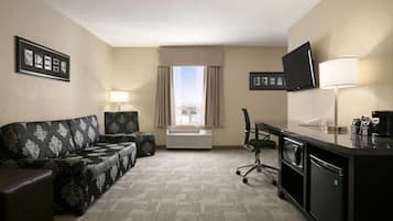 Habitación de hotel con sofá, escritorio, silla, televisión, lámpara y ventana con cortinas.