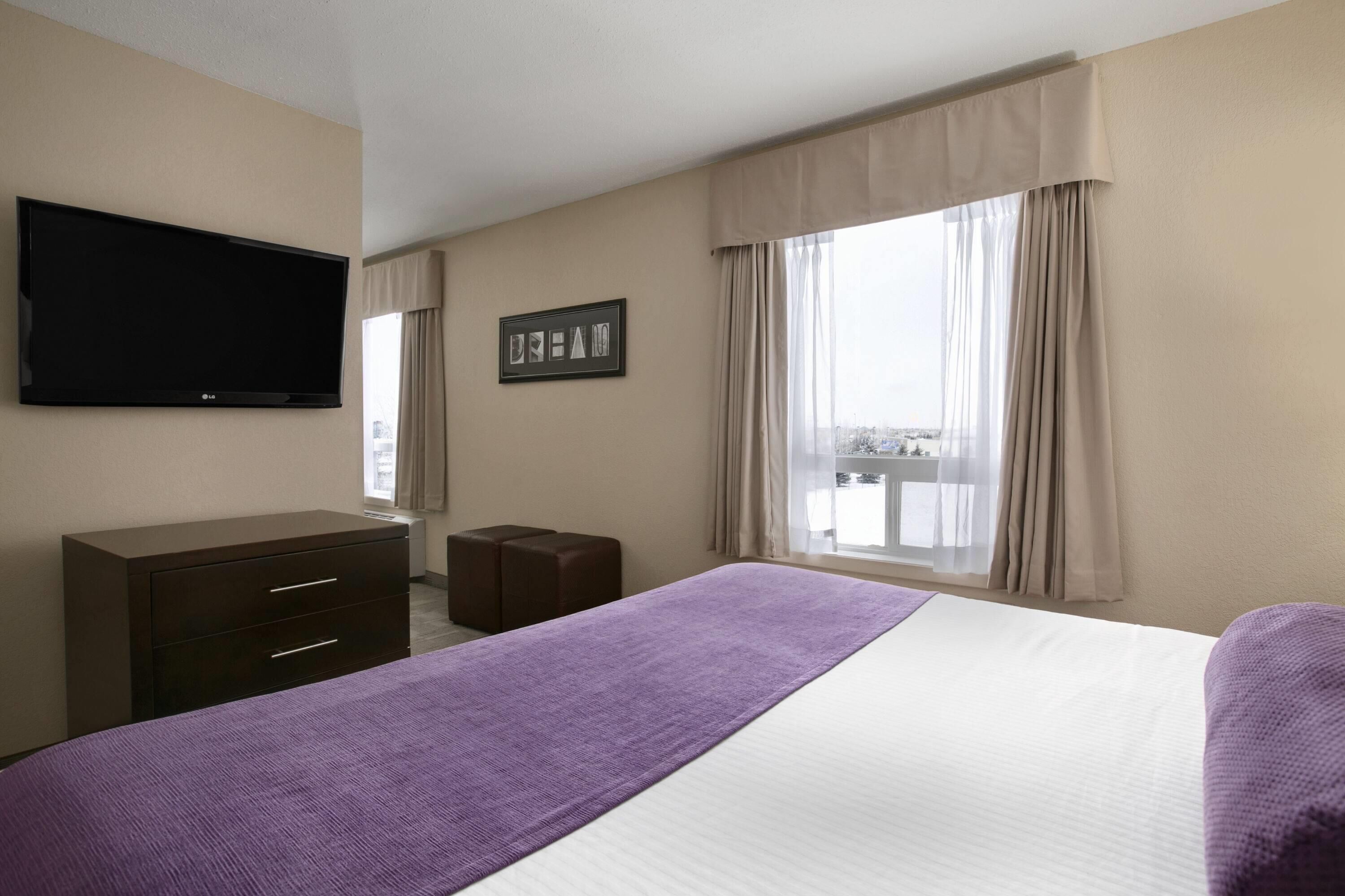A hotel room with a bed, a TV mounted on the wall, a small desk, and a window with curtains.
