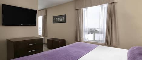 A hotel room with a bed, a TV mounted on the wall, a small desk, and a window with curtains.