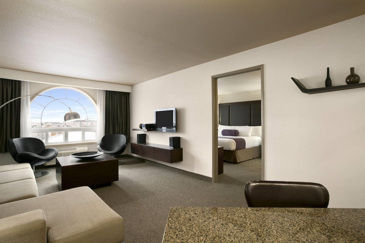 A modern hotel room with a large window, a flat-screen TV, a sofa, a coffee table, and a dining area.