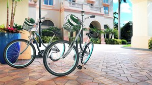 Two bicycles with baskets parked on a paved area in front of a building with balconies and arches.