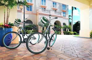 Two bicycles with baskets parked on a paved area in front of a building with balconies and arches.