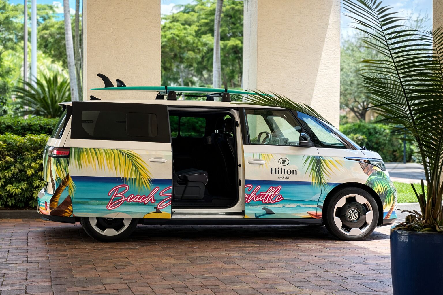 A beach-themed shuttle bus with 'Hilton Naples' branding, parked on a brick pavement with palm trees in the background.
