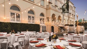 A formal outdoor dining area with round tables set for a meal, string lights overhead, and a building with arched windows in the background. - Hilton Naples (Naples)