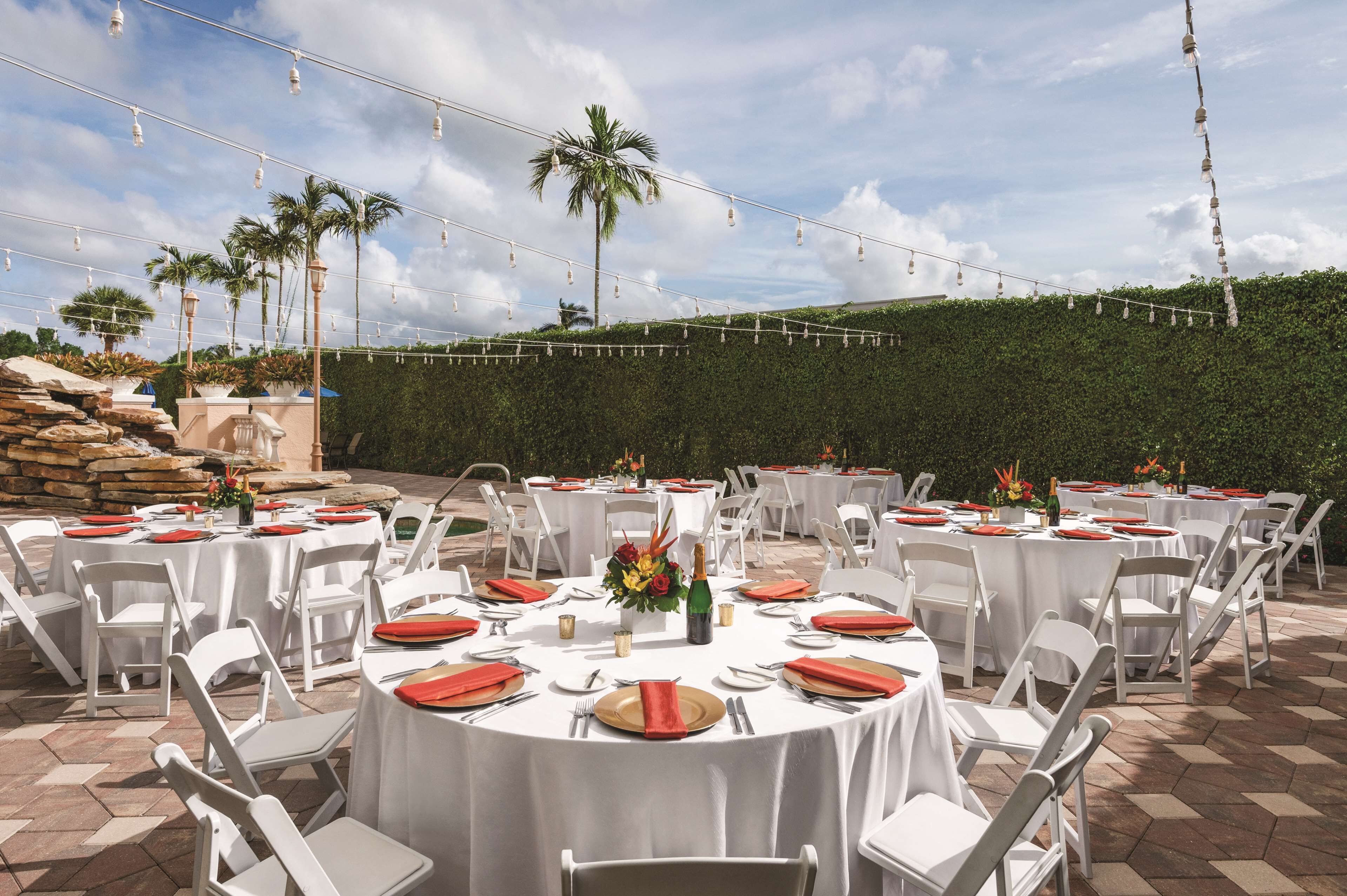 An outdoor dining area with round tables set for a formal event, surrounded by white chairs and a backdrop of palm trees and a hedge.