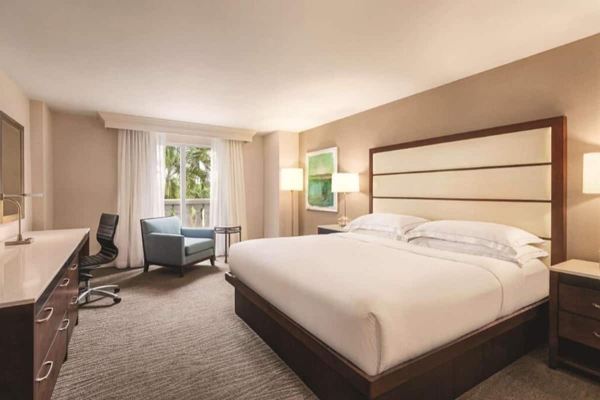 Une chambre d’hôtel avec un grand lit, un bureau avec une chaise, une petite table et une fenêtre avec des rideaux.