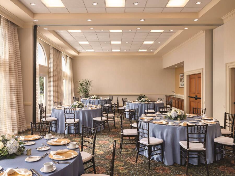 A banquet hall with round tables set for dining, featuring floral centerpieces and a carpeted floor.